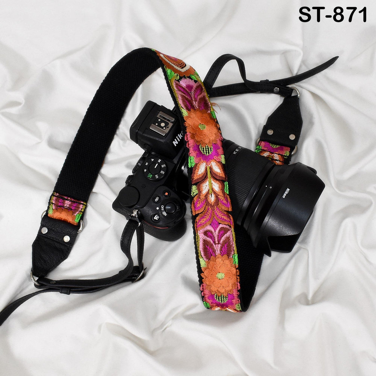 A black camera strap with a colorful floral pattern, a camera lens, and a camera body are arranged on a white background.