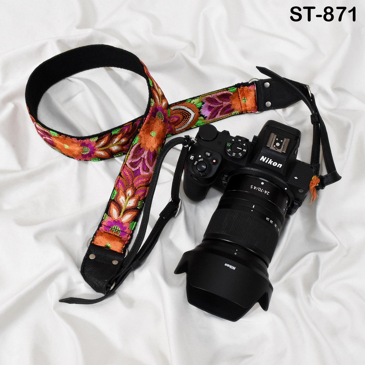 A black and orange floral camera strap and a Nikon camera with a lens attached are placed on a white surface.