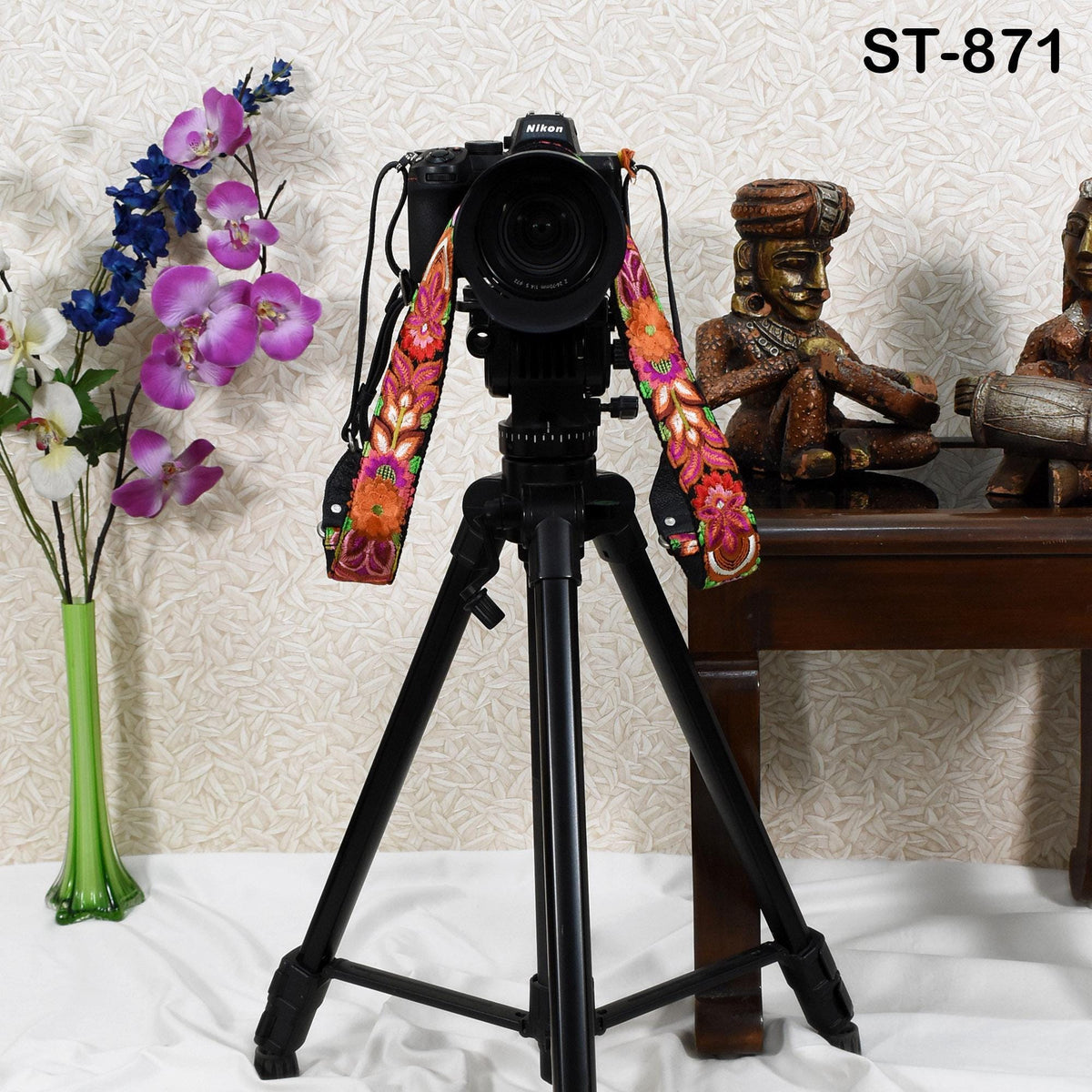 A black tripod with a camera attached to it, along with a colorful strap, is placed on a table next to a vase of purple flowers and a figurine.