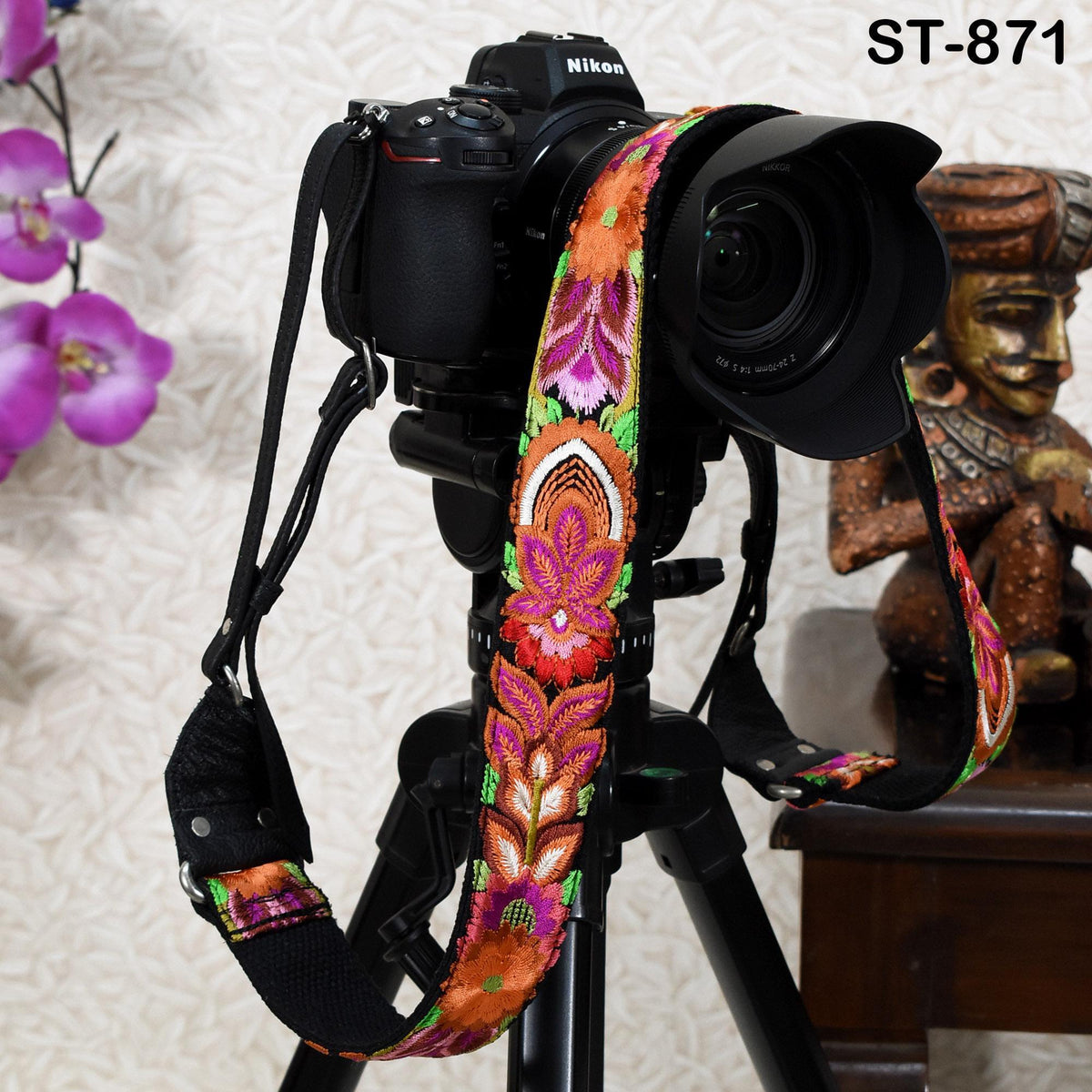 A black camera with a colorful floral patterned strap is mounted on a tripod, with a wooden statue visible in the background.