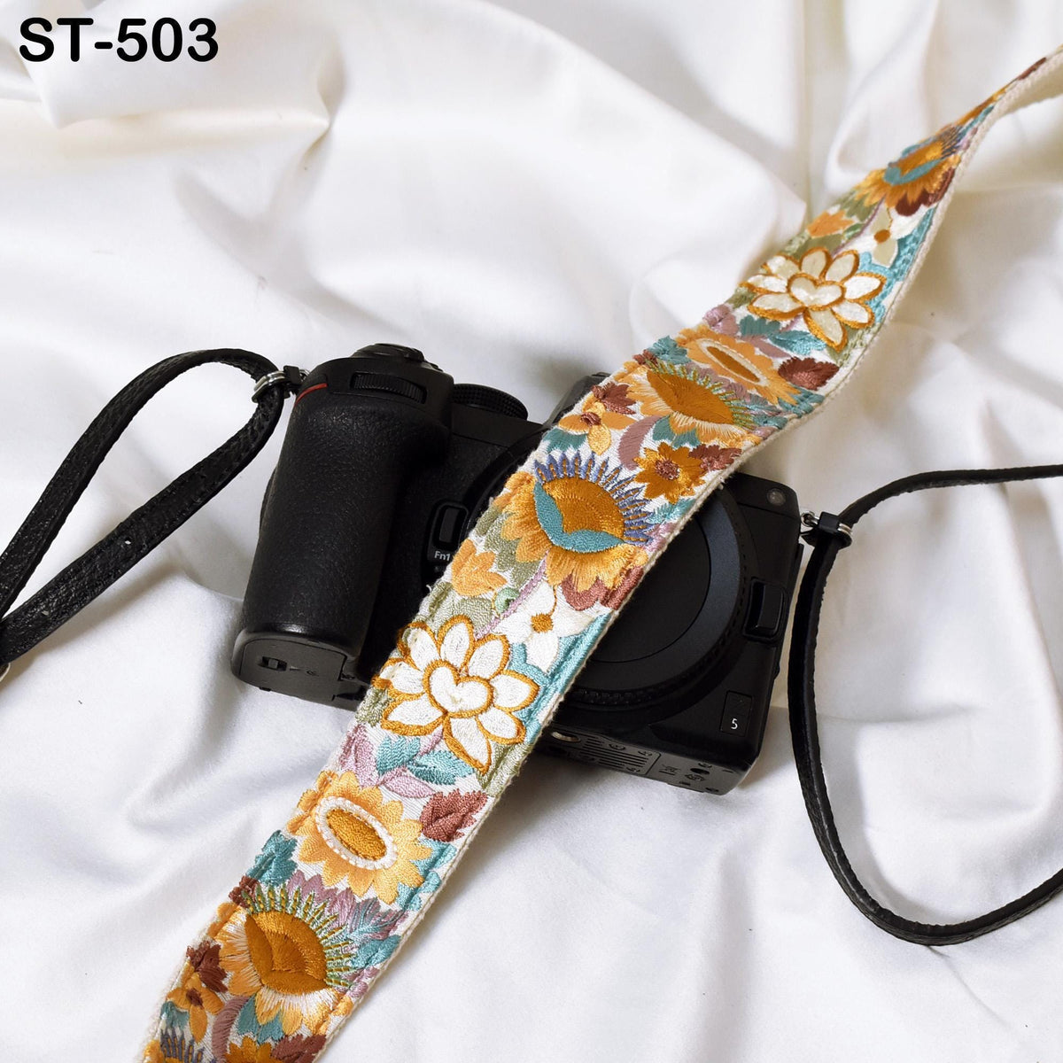 A black camera strap with a colorful floral pattern is draped over a camera, resting on a white fabric surface.