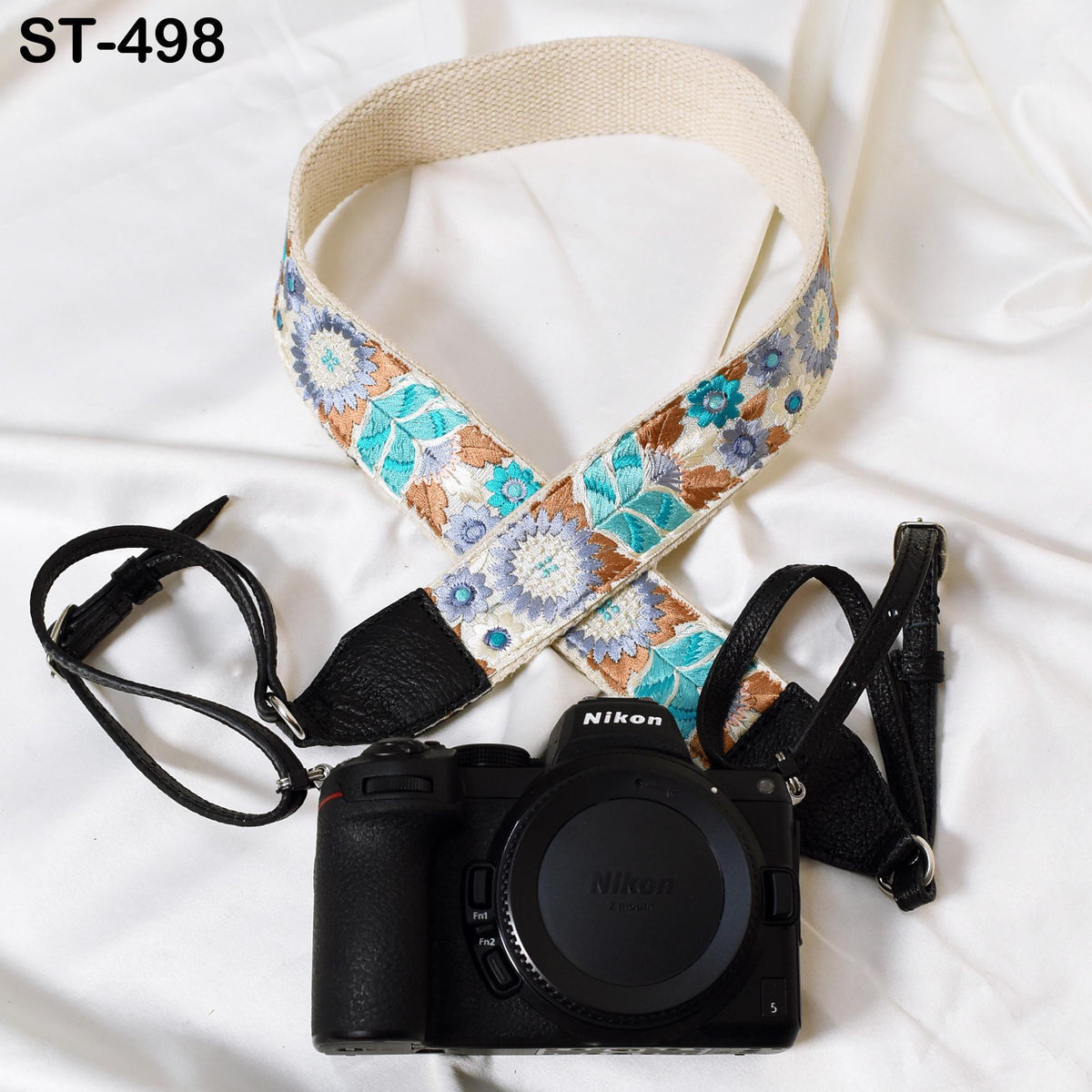 A black camera with a floral patterned strap and a white strap are placed on a white surface.