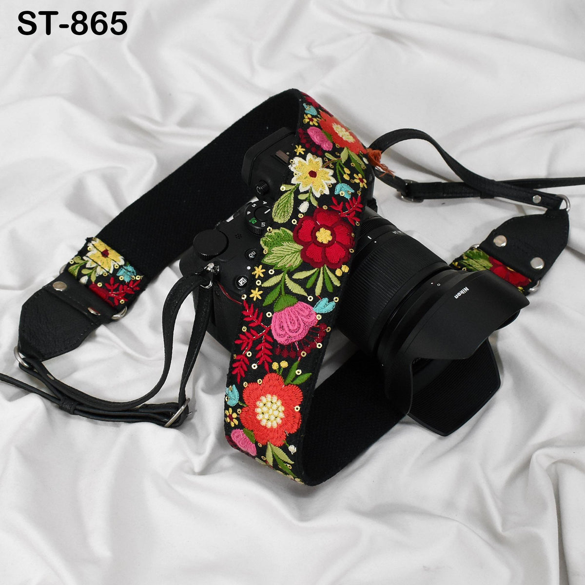 A floral embroidered camera strap is draped over a camera lens, resting on a white fabric surface.