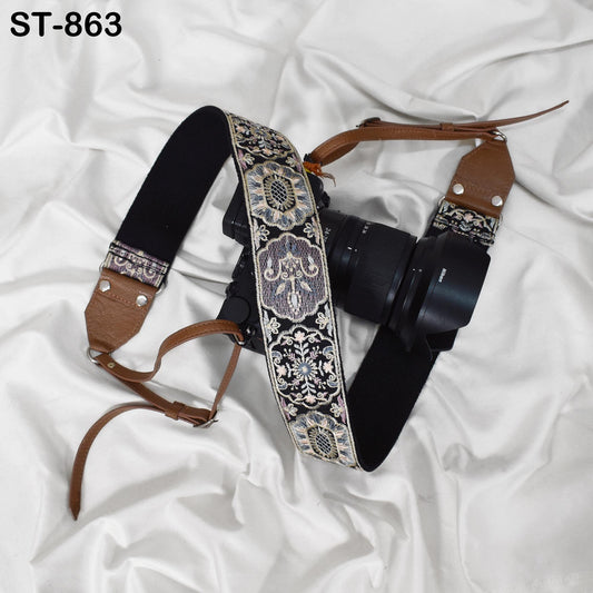 A camera strap with a decorative pattern and a camera lens attached to it, both resting on a white fabric surface.