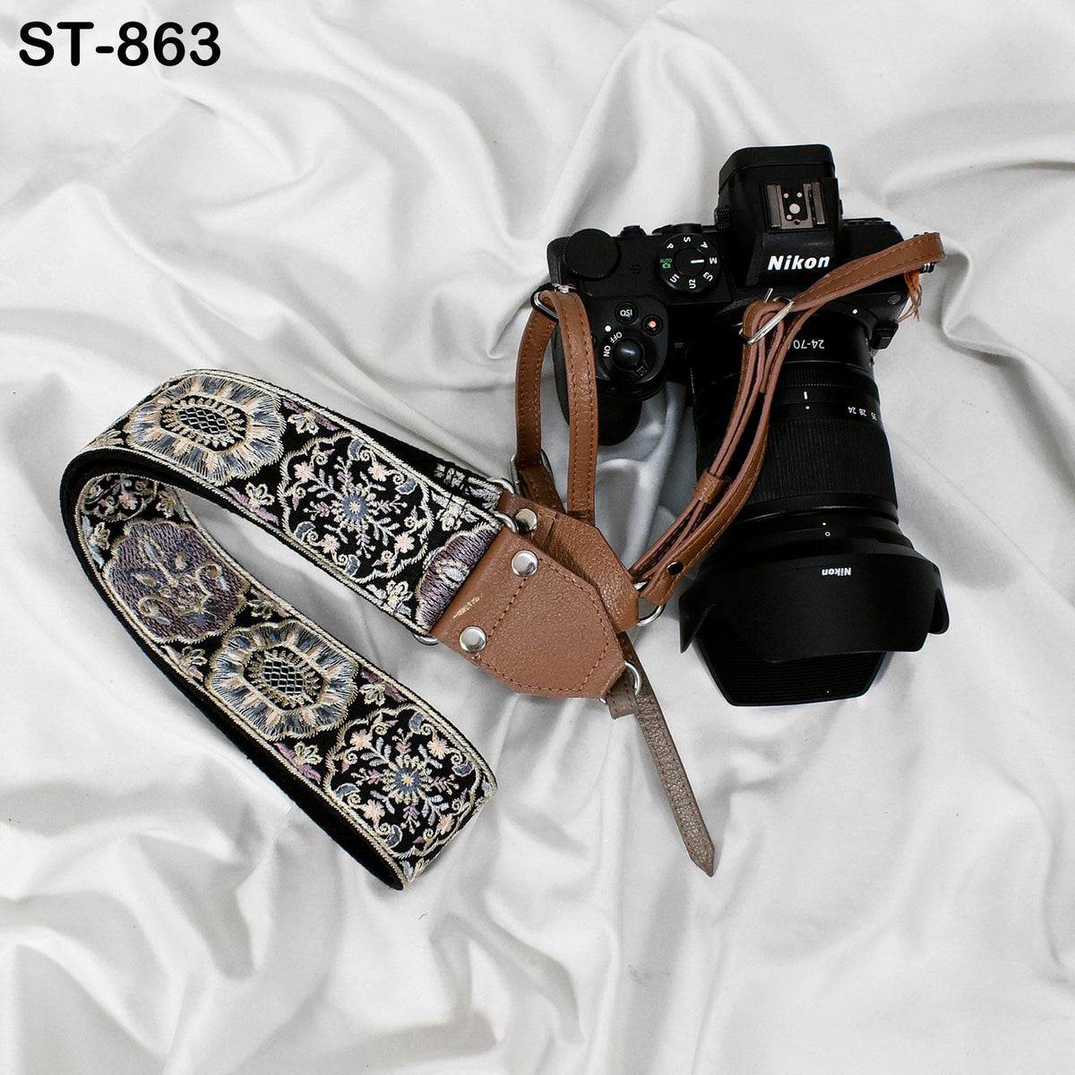 A black and brown camera strap with a floral pattern and a Nikon camera body are placed on a white fabric surface.
