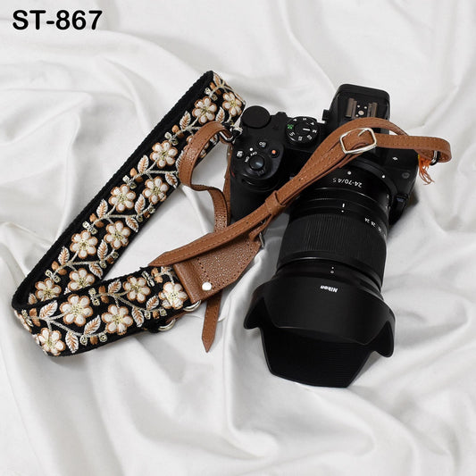 A black and brown camera strap with a floral pattern is attached to a black camera lens, resting on a white surface.