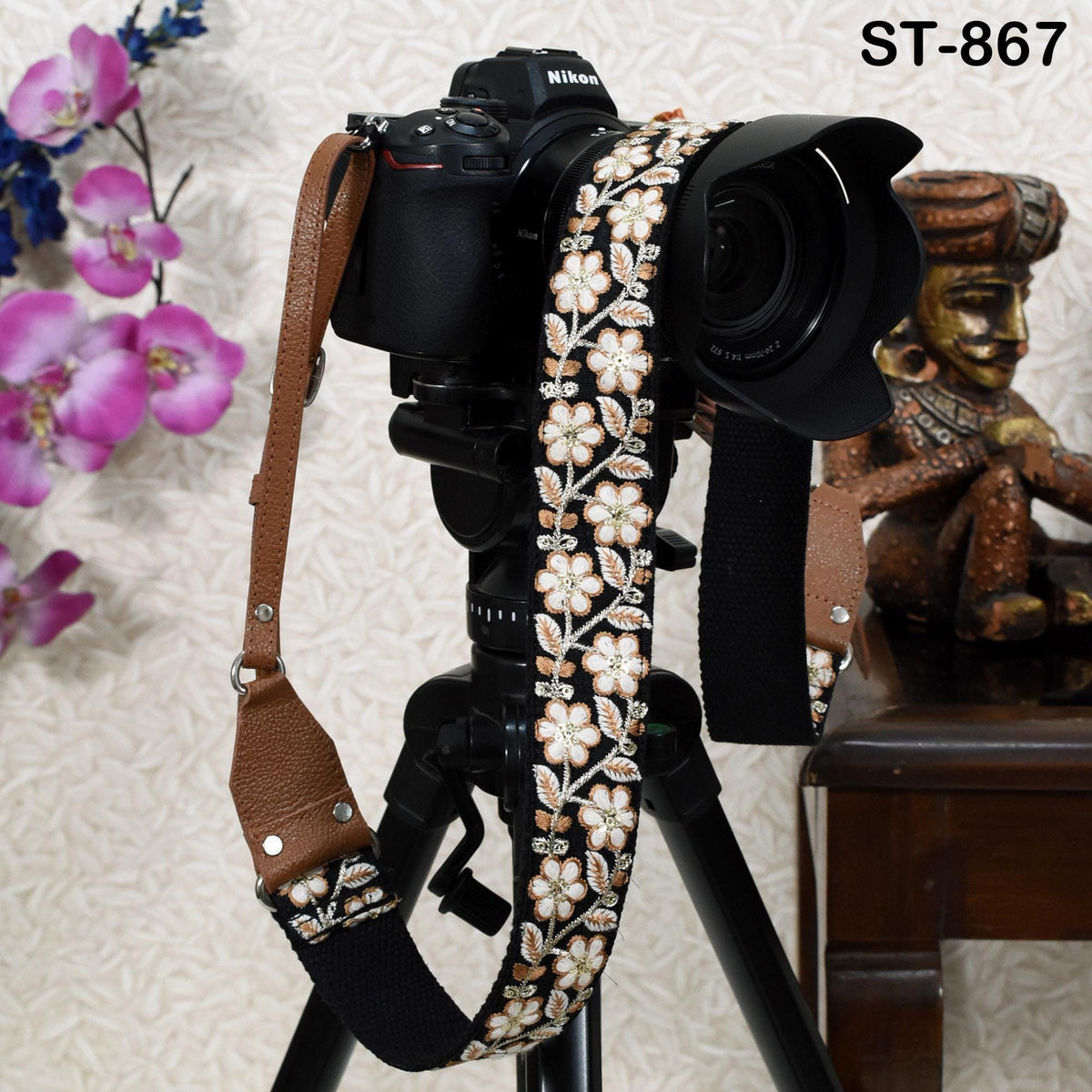 A black camera with a floral patterned strap is mounted on a tripod, with a wooden table and a statue in the background.