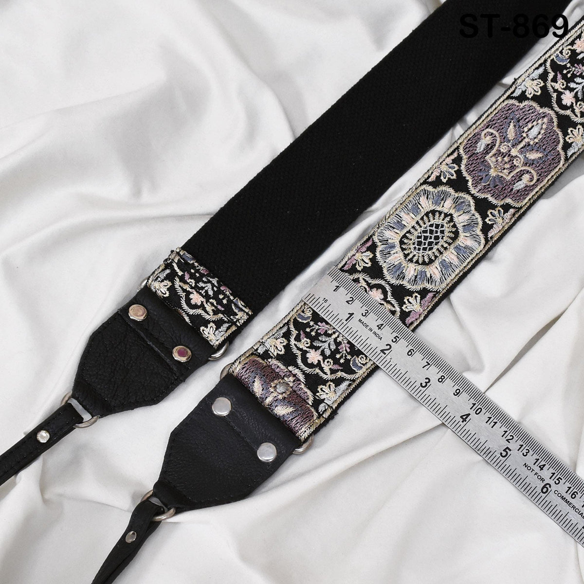 A black and silver camera strap with intricate embroidered patterns, resting on a white fabric surface.