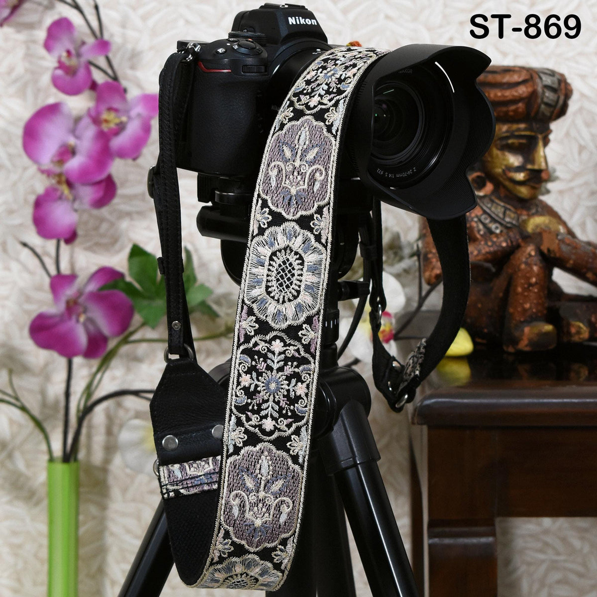 A black camera with a decorative patterned strap is placed on a tripod, with a wooden statue and purple orchids in the background.