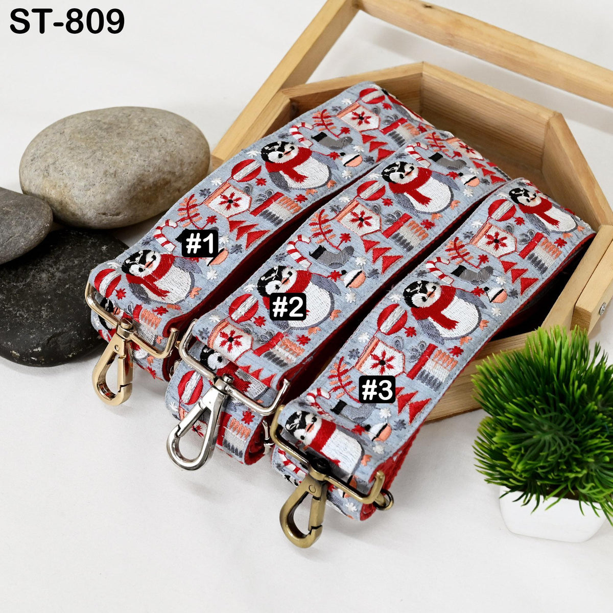 a wooden box containing three colorful straps with a snowman pattern, placed on a white surface alongside some rocks and a potted plant.