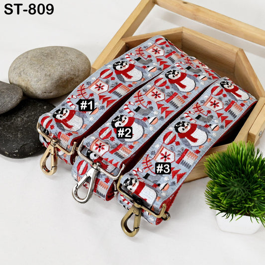 a wooden box containing three colorful straps with a snowman pattern, placed on a white surface alongside some rocks and a potted plant.
