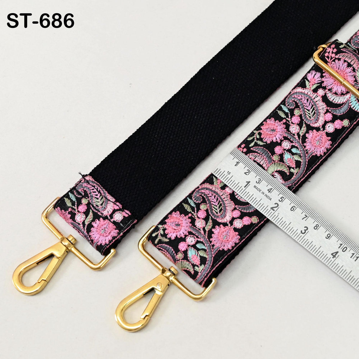 A black strap with a floral pattern and two gold metal hooks, along with a ruler for size reference.