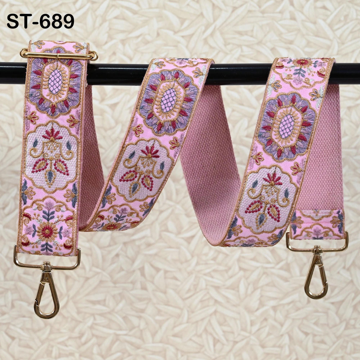 A pink floral embroidered strap with gold hardware, hanging on a black rod.