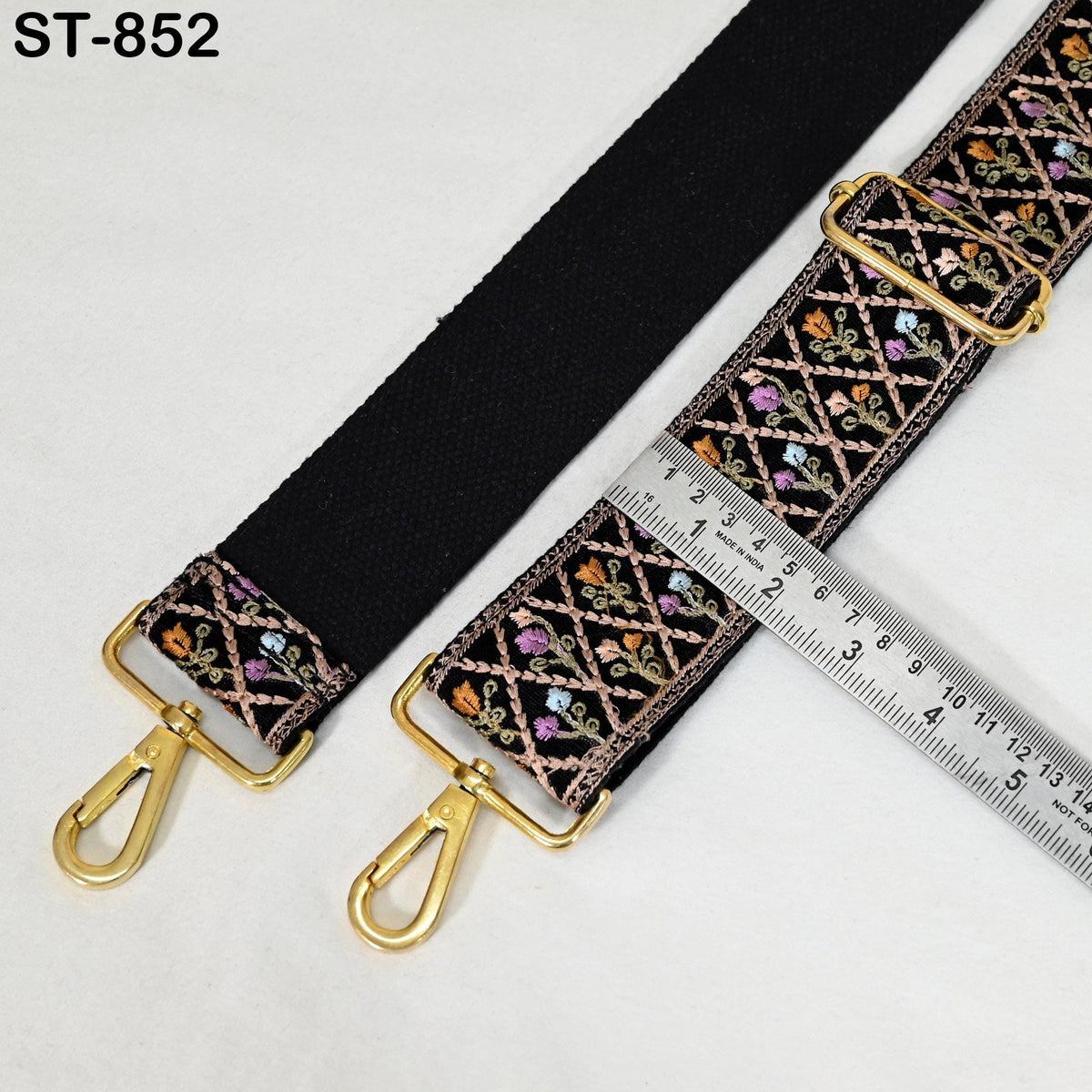 a black strap with a floral pattern and two gold metal hooks, placed next to a ruler for size comparison.