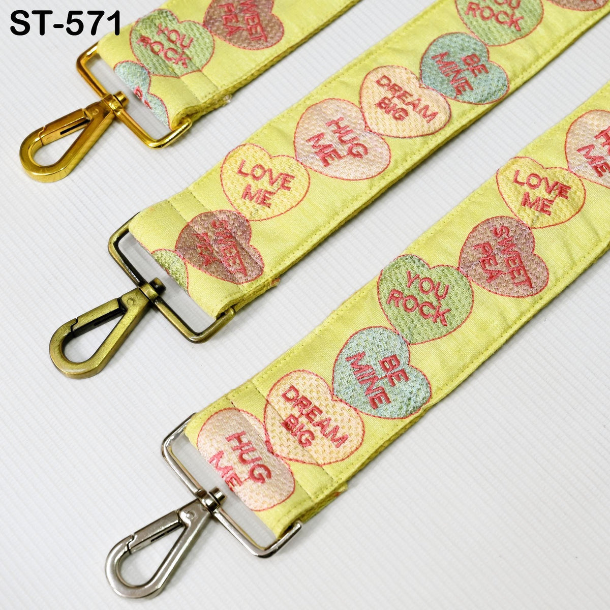 a yellow strap with a heart-shaped design and various phrases printed on it, such as "LOVE ME", "HUG ME", "DREAM BIG", "SWEET REY", "BE MINE", and "SWEET SWEET". The strap has a metal hook at the top and a metal loop at the bottom, suggesting it is designed for attaching or detaching from a bag or other accessory.