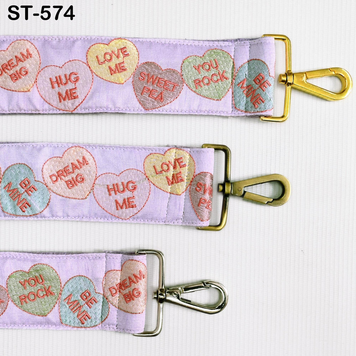 a purple strap with heart-shaped embroidered phrases such as "HUG ME", "LOVE ME", "SWEET PEA", "BE ME", and "DREAM BIG". The strap has a gold metal clasp and a silver metal loop, and the strap is displayed against a white background.