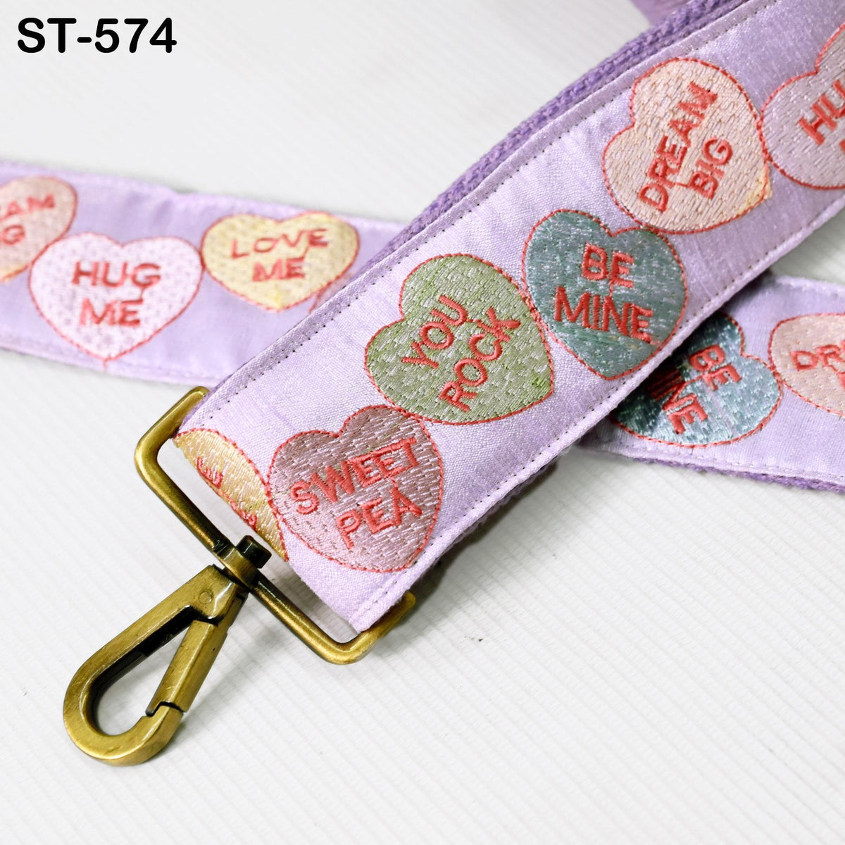 A purple strap with heart-shaped embroidered phrases such as "HUG ME", "LOVE ME", "BE MINE", "YOU ROCK", "SWEET PEAS", and "DREAM BIG" on it. The strap also has a gold metal clasp.