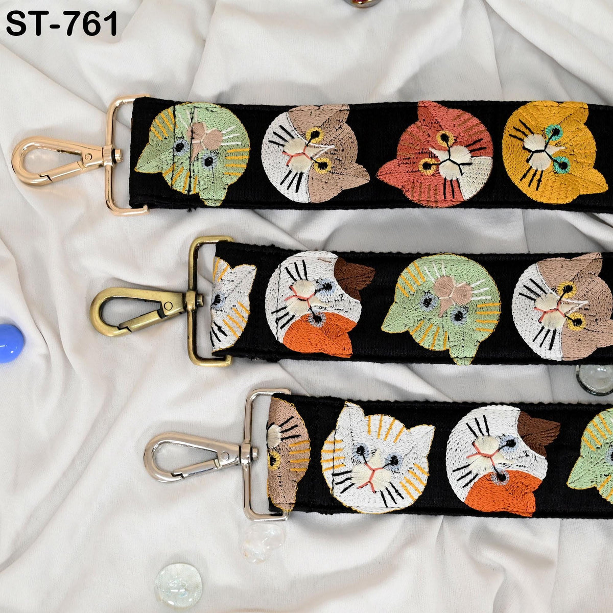 a black strap with a repeating pattern of cat faces in various colors, including green, orange, and yellow. The strap has a gold buckle and a silver hook, and there are small objects scattered around the strap.