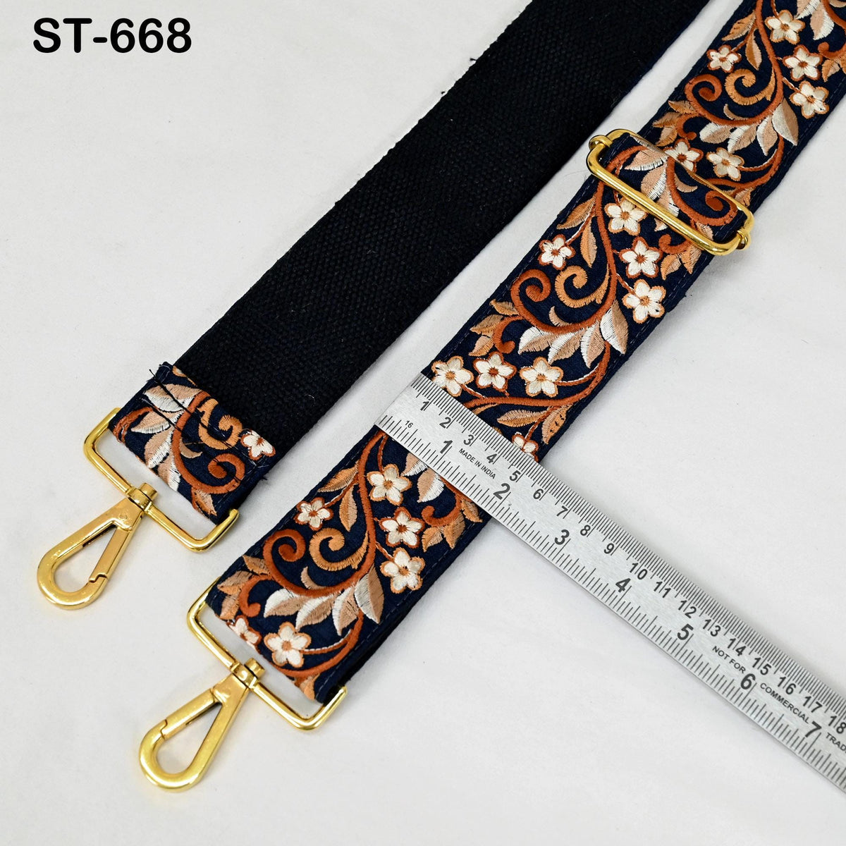 a black strap with a floral pattern and two gold metal hooks, placed next to a ruler for size comparison.