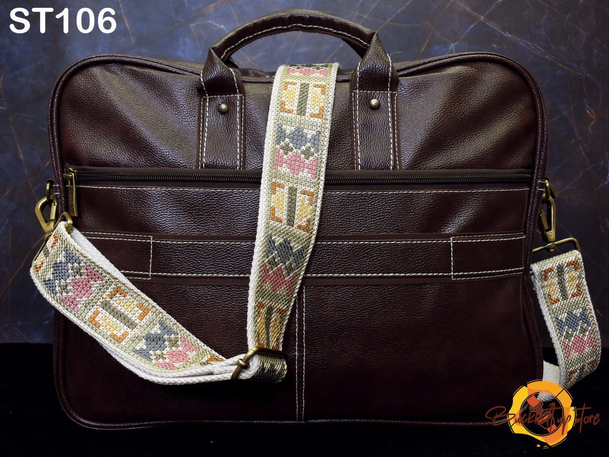 A brown leather laptop bag with a cross-stitch patterned strap, displayed against a dark background.