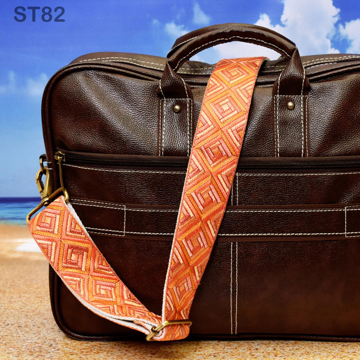 A brown leather briefcase with a patterned strap is placed on a sandy beach, with a clear blue sky and ocean in the background.