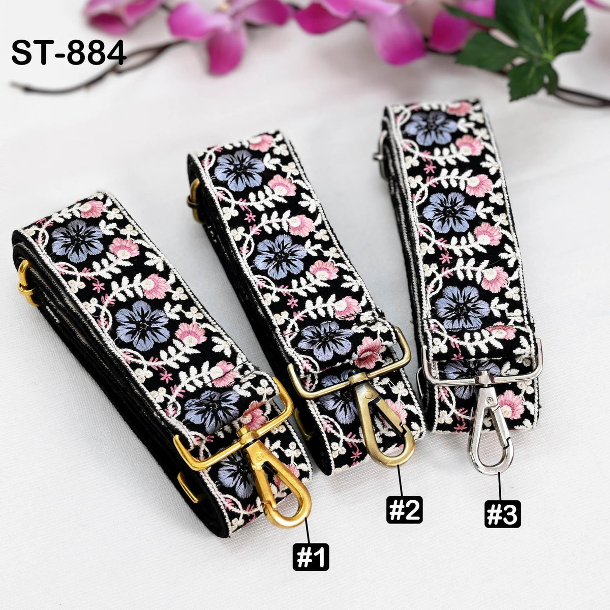 Three floral-patterned straps with gold buckles, each with a different number printed on it, placed on a white surface with pink flowers in the background.