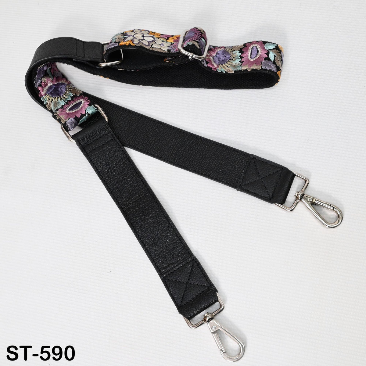 A black strap with a floral pattern and metal hooks attached to it, lying on a white surface.